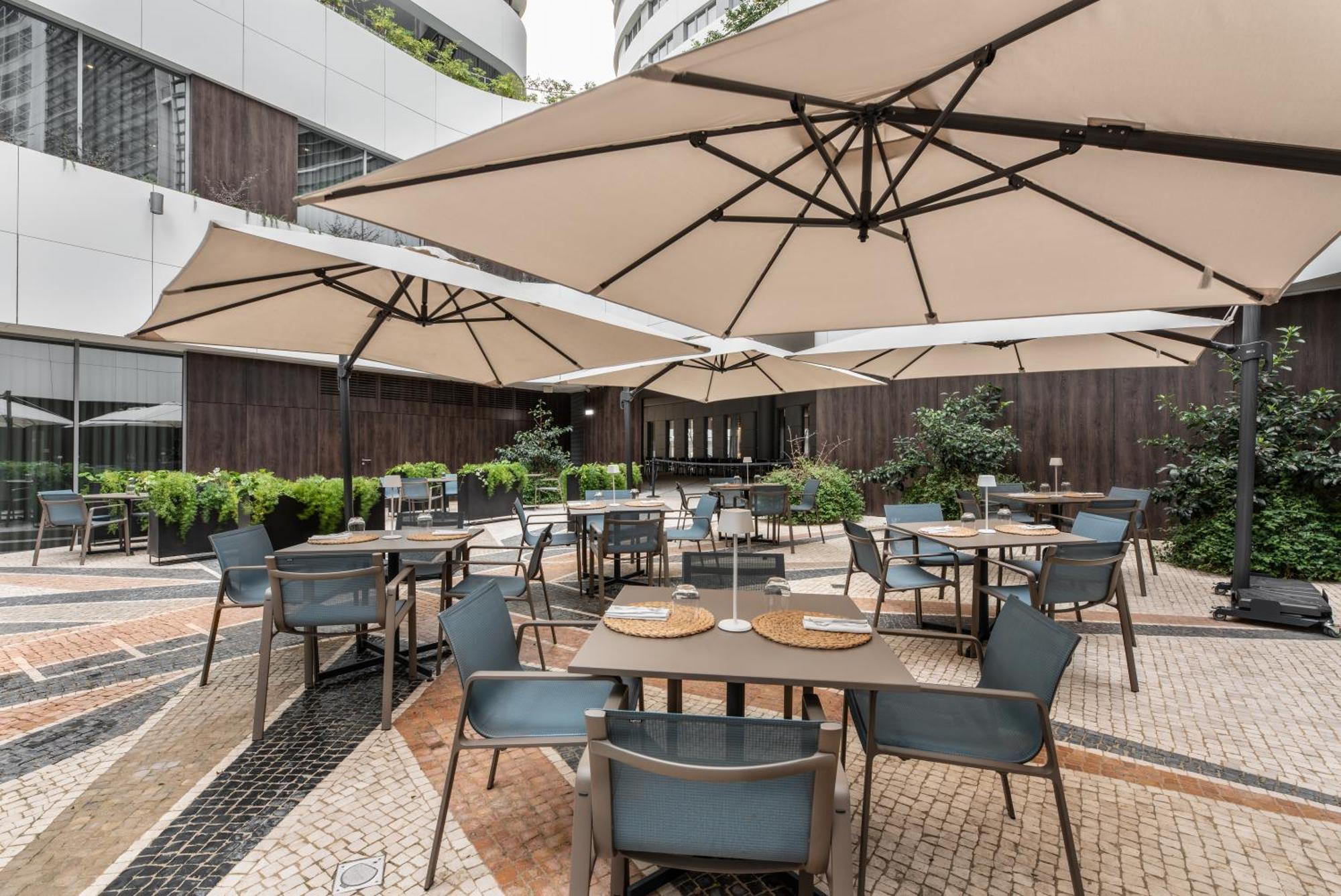 Eurostars Universal Lisboa 外观 照片 The photo shows an outdoor dining area featuring several tables and chairs set up beneath large umbrellas. The space is designed with a modern aesthetic, surrounded by greenery and potted plants. The flooring consists of patterned tiles, adding to th