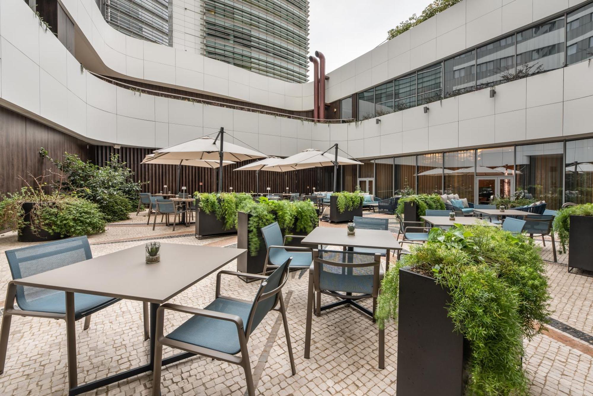 Eurostars Universal Lisboa 外观 照片 The image shows a modern outdoor seating area, likely part of a restaurant or cafe. It features several tables and chairs arranged neatly, with some tables covered by umbrellas for shade. Lush greenery surrounds the area, with planters filled with pl