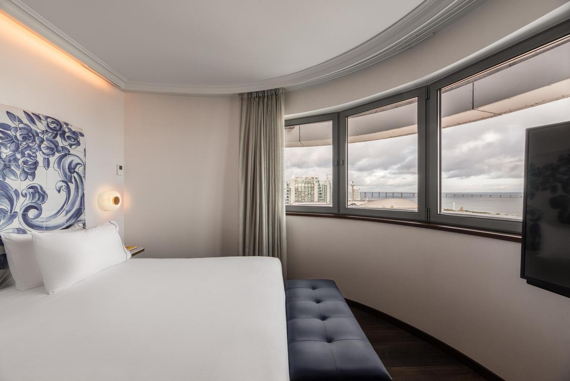 Eurostars Universal Lisboa 外观 照片 The photo shows a modern hotel room with a curved wall design. It features a bed with white linens positioned against the wall. There is a stylish ottoman or bench at the foot of the bed. The room has large windows that provide a view, allowing natur