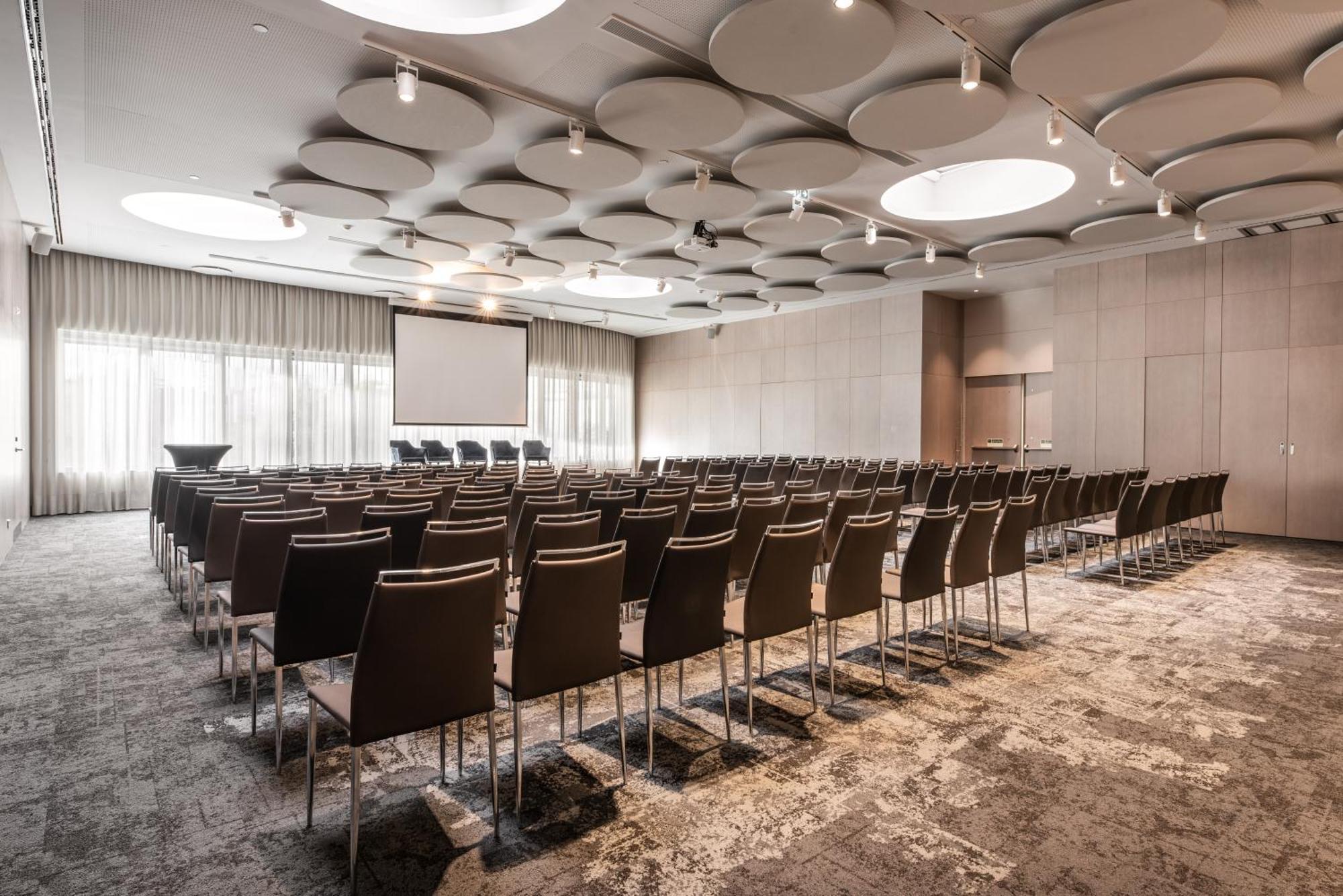 Eurostars Universal Lisboa 外观 照片 The photo shows a conference or meeting room that is set up for an event. There are rows of brown chairs arranged facing a screen at the front of the room. The space has a modern design, with a decorative ceiling featuring circular elements. Large wi