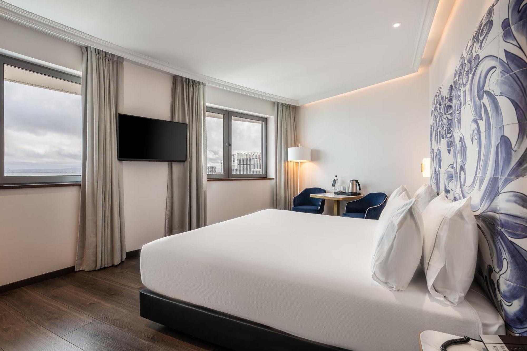 Eurostars Universal Lisboa 外观 照片 The photo shows a modern hotel room. It features a large bed with white linens, a small table with two blue chairs, and a flat-screen television mounted on the wall. There are windows with light-colored curtains that let in natural light, and a decor