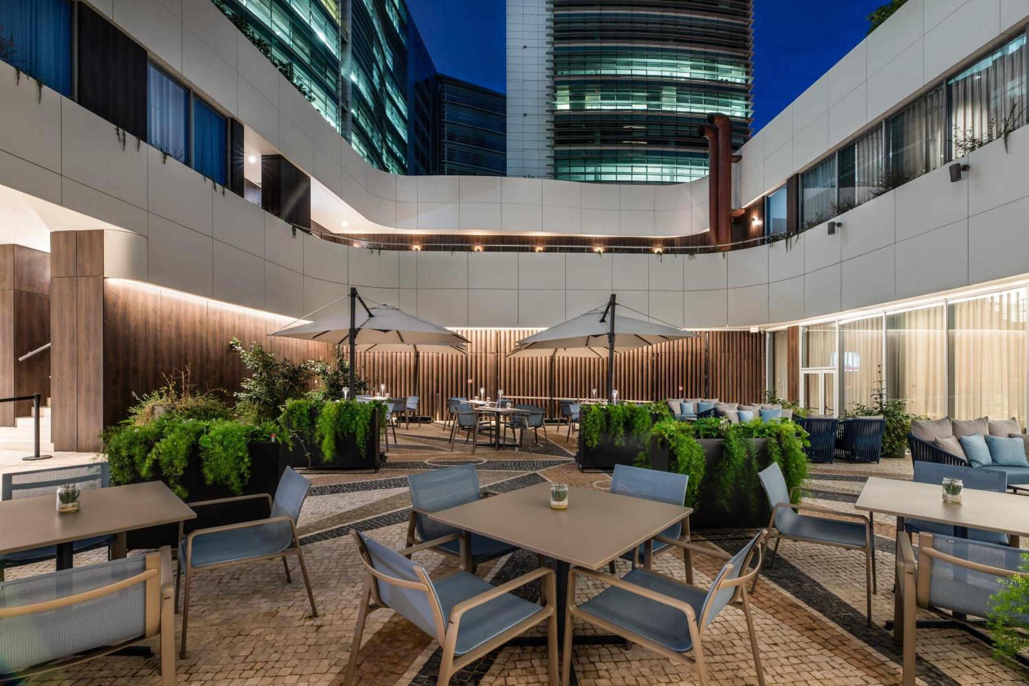 Eurostars Universal Lisboa 外观 照片 The photo depicts an outdoor seating area likely within a modern urban setting, possibly a hotel or office complex. It features several tables and chairs arranged around the space, with large umbrellas providing shade. Lush greenery is seen in variou